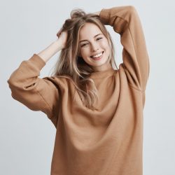 Beautiful pretty charming young blonde woman in loose sweater smiling happily, having fun indoors, playing with long straight hair. Pretty girl looking at camera with joyful smile, rejoicing life