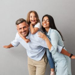 Portrait of a cheerful family father, mother, little daughter having fun together, father holding little daughter on his back isolated over gray background