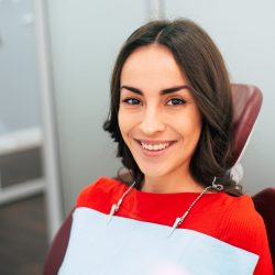 Dental care. Shining girl is smiling with her stunning teeth and sitting in the dental chair. She is wearing a red sweater and there is also a protective cloth on her neck.