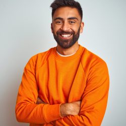 Young indian man wearing orange sweater over isolated white background happy face smiling with crossed arms looking at the camera. Positive person.