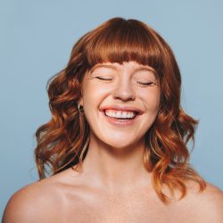 Happy woman with ginger hair and flawless skin smiling with her eyes closed. Cheerful young woman embracing her natural beauty. Body confident young woman standing against a blue studio background.
